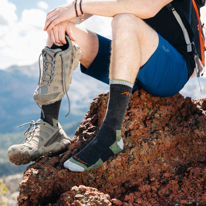 Full shot of model sitting on edge of a mountain wearing the men's boot hiking sock in lime and putting on taupe hiking shoes