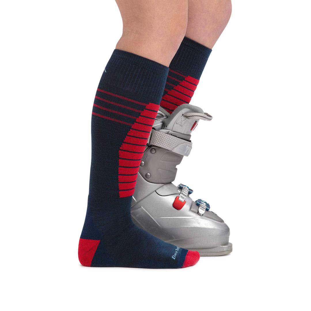 Kid wearing Edge Over-the-Calf Ski & Snowboard socks in Eclipse with red design on shin with back foot in a ski boot to see height of sock, Lifestyle Image