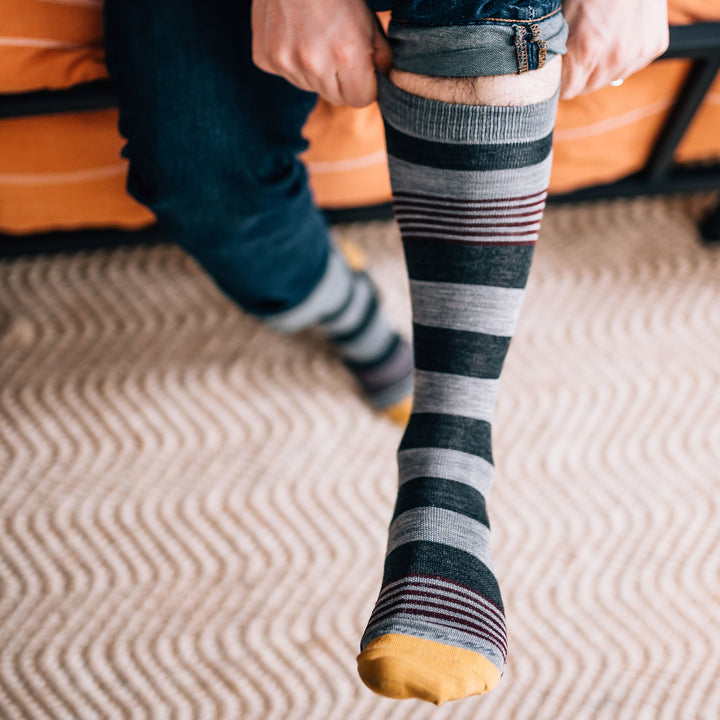 Man sitting on a couch, jeans rolled up pulling on his Oxford Crew Lightweight Lifestyle Socks in Gray