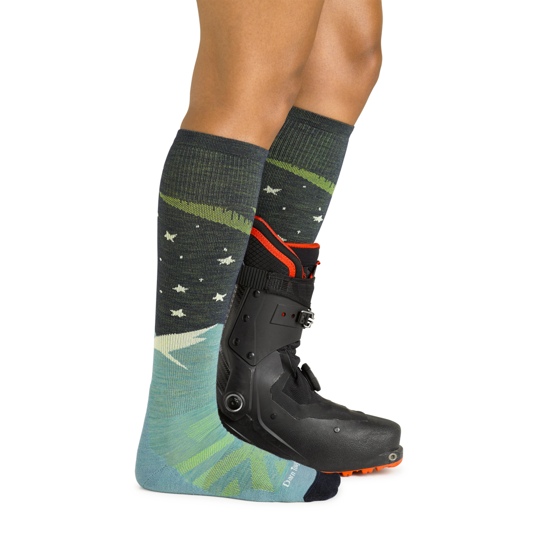 Model wearing women's aurora over-the-calf lightweight snow sock in aqua with black ski boot on left foot