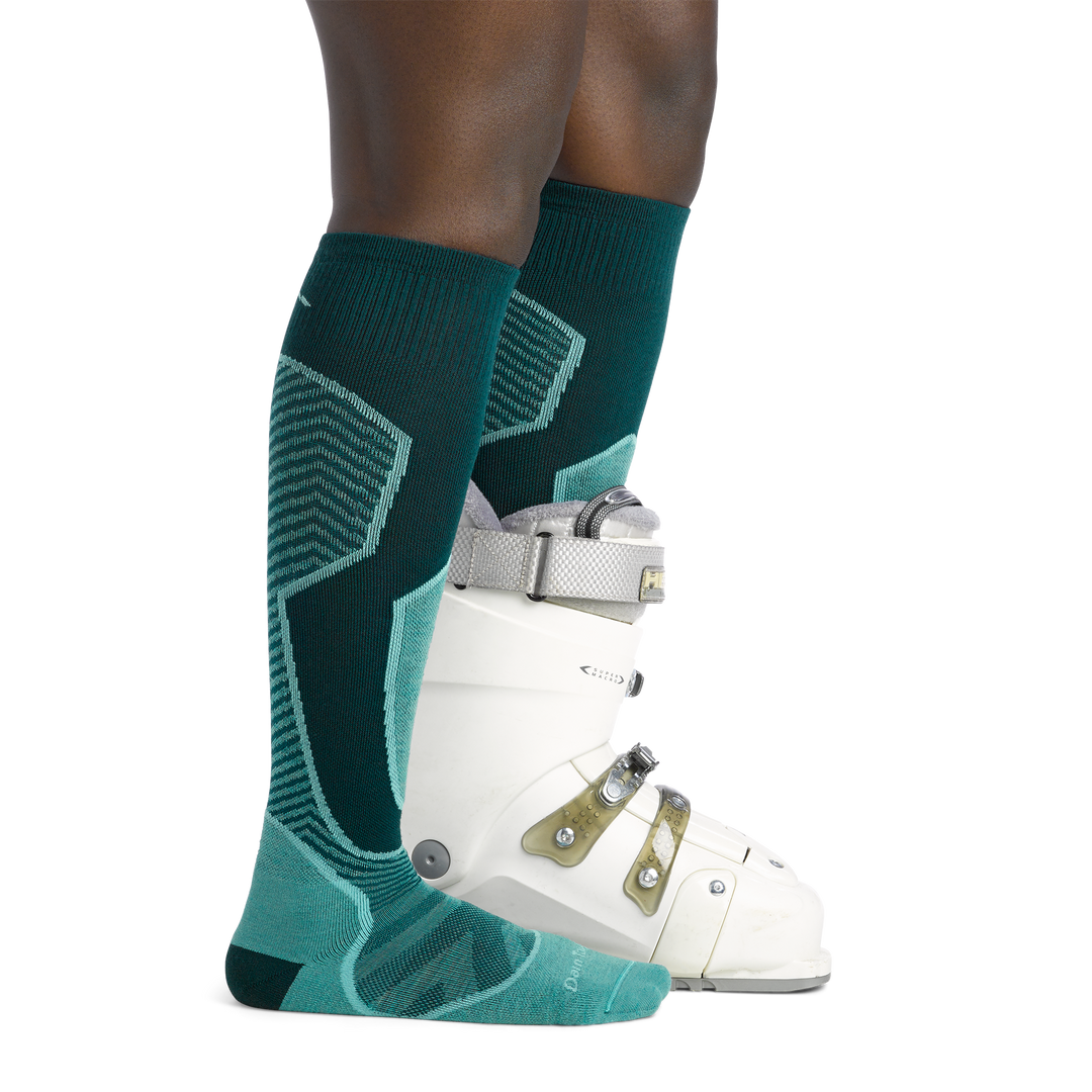 Woman facing to the right, wearing Outer Limits Over the Calf Ski & Snowboard Socks in Juniper, back foot also wearing a snowboard boot