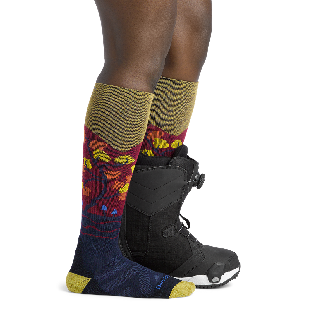 Woman facing to the right, wearing Enchanted Over the Calf Ski & Snowboard Socks in Autumn, back foot wearing a snowboard boot