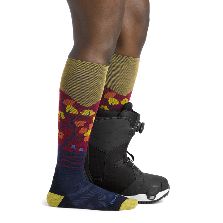 Woman facing to the right, wearing Enchanted Over the Calf Ski & Snowboard Socks in Autumn, back foot wearing a snowboard boot