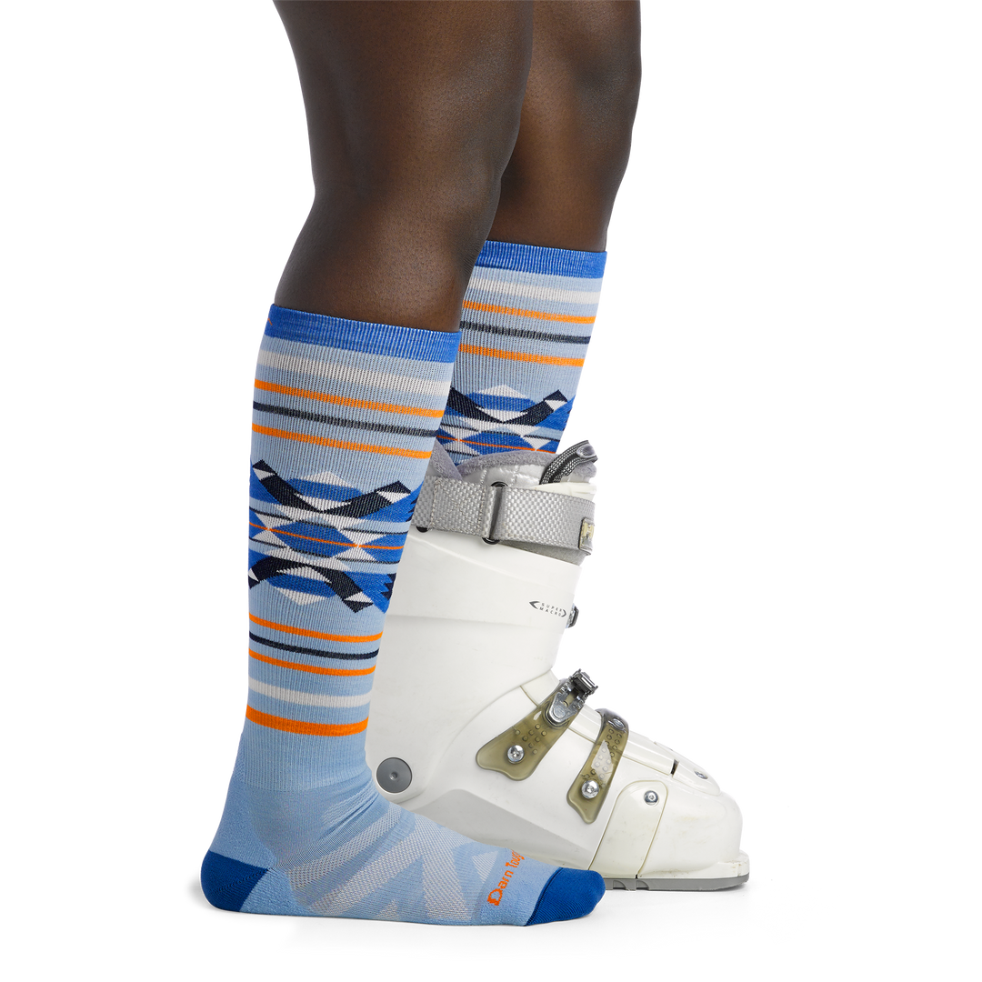 Woman facing to the right, wearing Snowscape Over the Calf Ski & Snowboard Socks in Bluebird, back foot wearing a snowboard boot