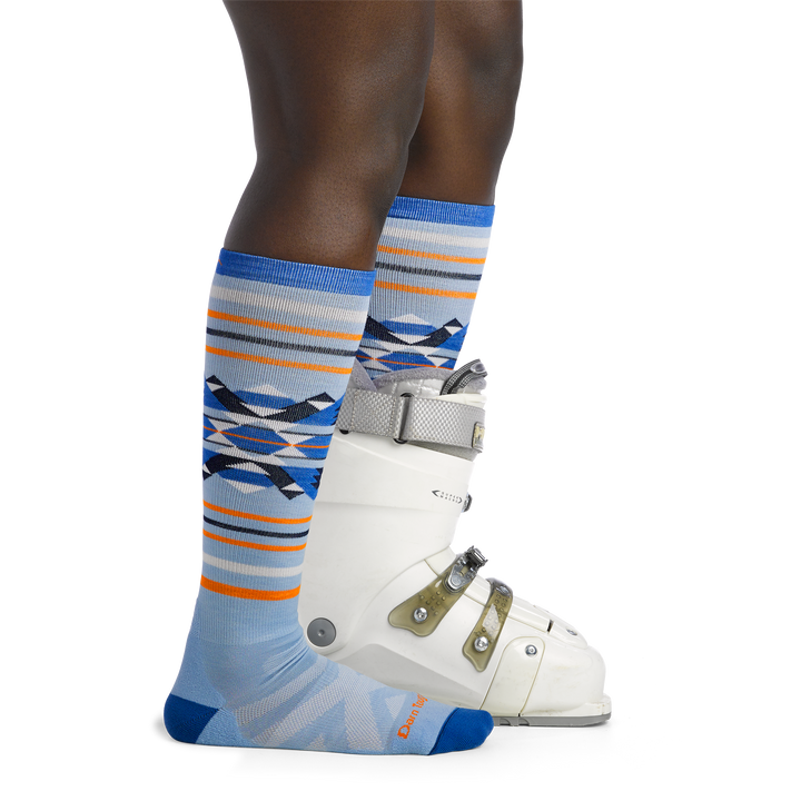 Woman facing to the right, wearing Snowscape Over the Calf Ski & Snowboard Socks in Bluebird, back foot wearing a snowboard boot