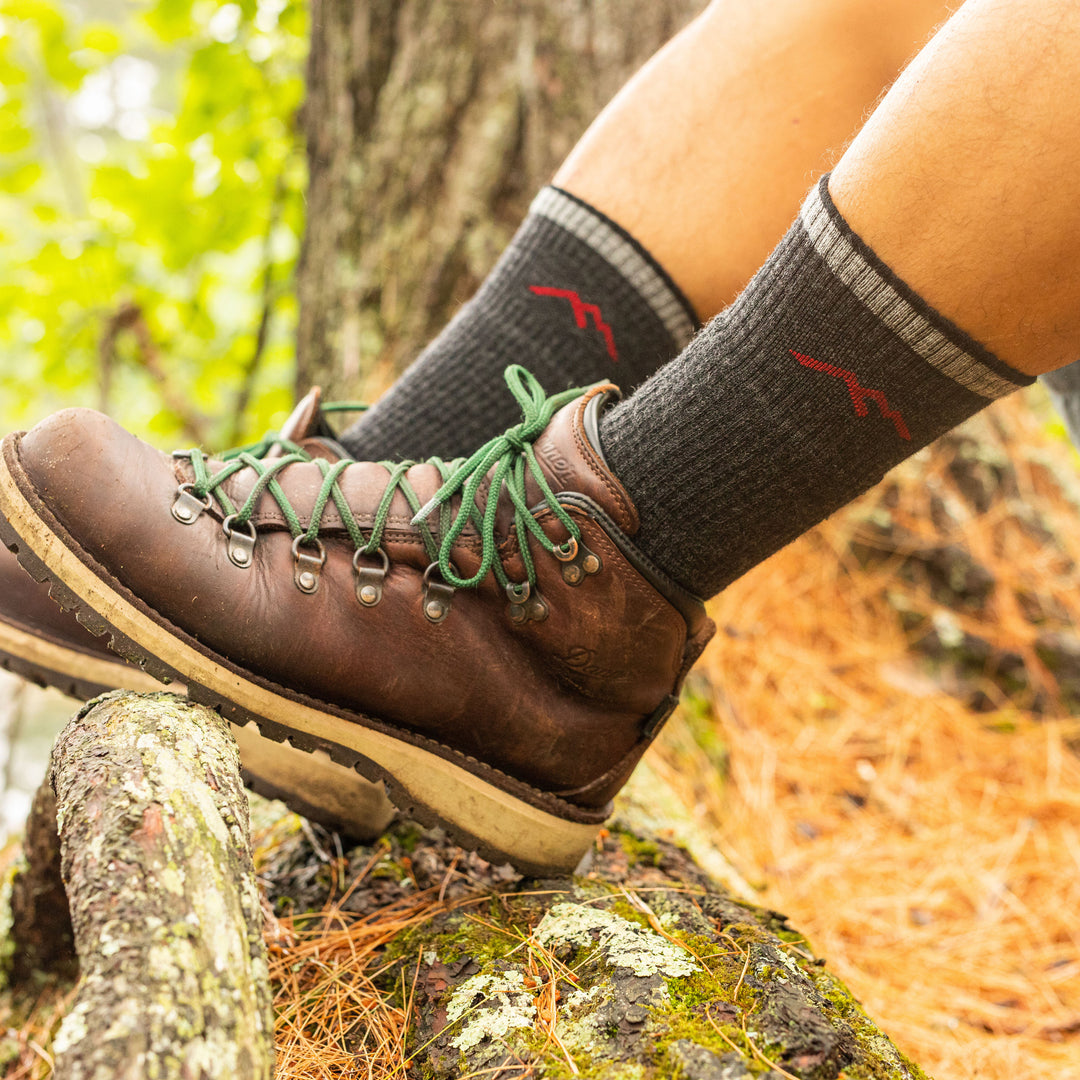 Side shot of model sitting on a rock wearing the men's boot hiking sock in black and dbrown hiking boots with green laces