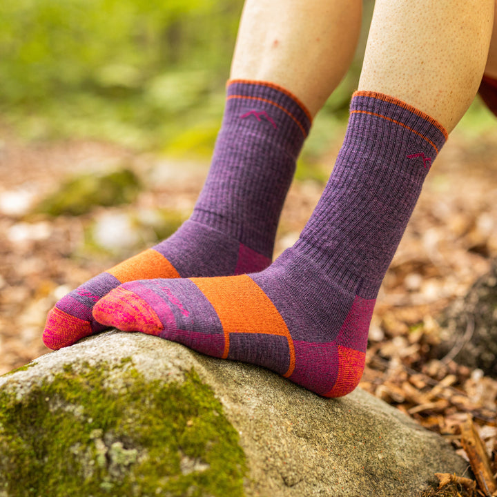 Close up lifestyle shot of model wearing hiker boot socks in Plum Heather, resting feet on a rock in the woods.