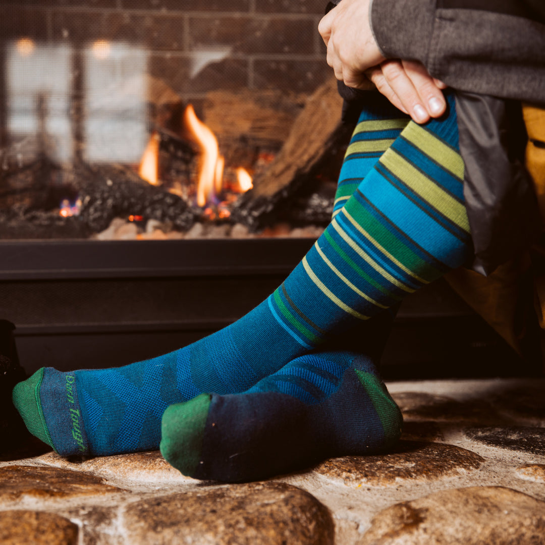 Close up side of model sitting on the ledge of a fireplace wearing the men's snowpack over-the-calf snow sock in midnight