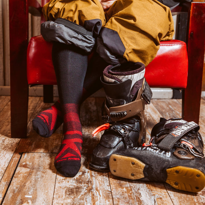 Close up shot of model sitting in a red chair in snowboard gear wearing the men's thermolite over-the-calf snow sock in black