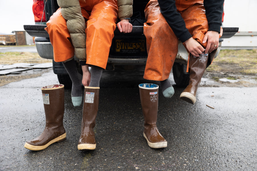 Two people seated on tailgate pulling on rubber boots over darn tough socks for work boots