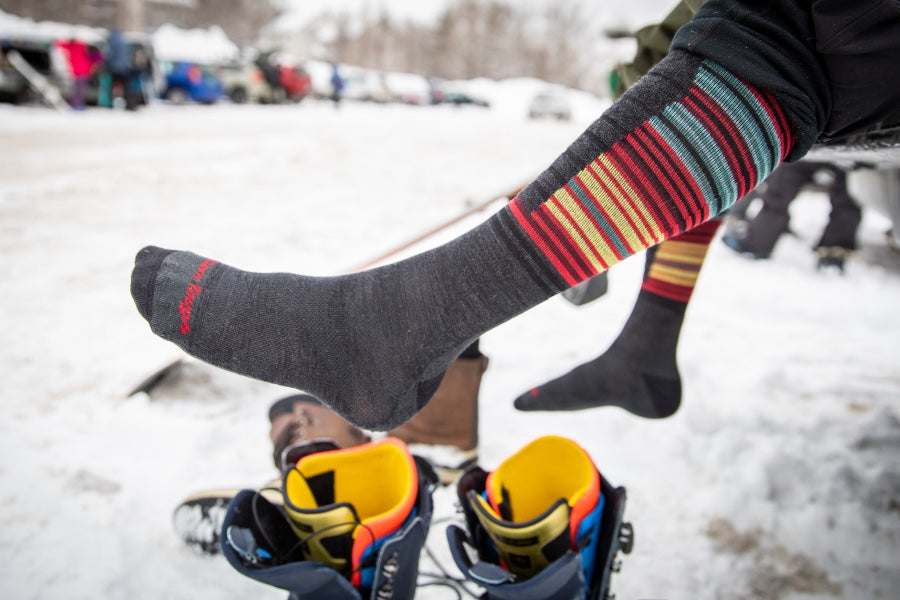 A skier about to put on ski boots wearing the Backwoods ski and snowboard sock with stripes