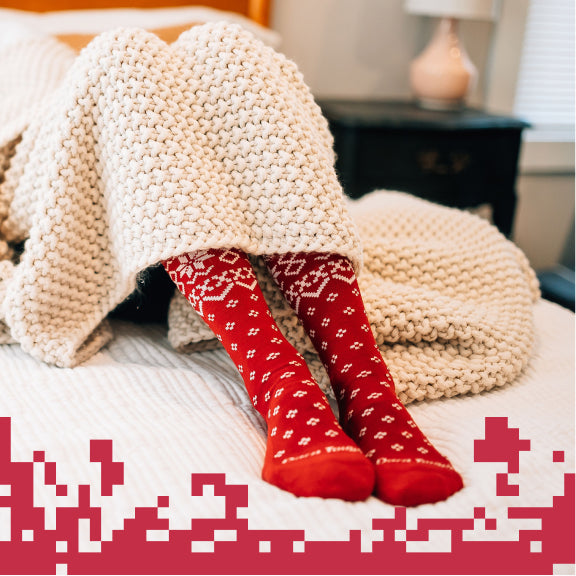 Feet sticking out from under a blanket looking cozy in red socks with a holiday-inspired pattern