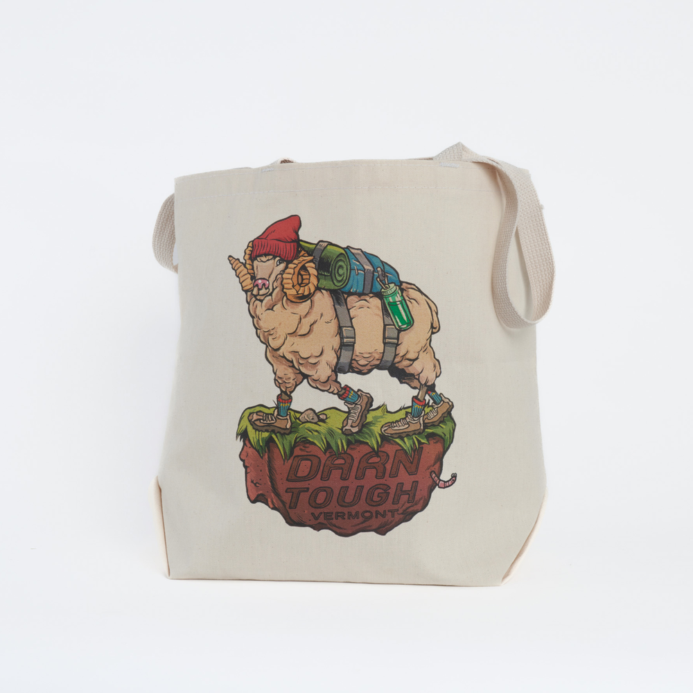 canvas tote bag with straps hanging , front of bag features art work of a northern ram wearing a hat and back pack