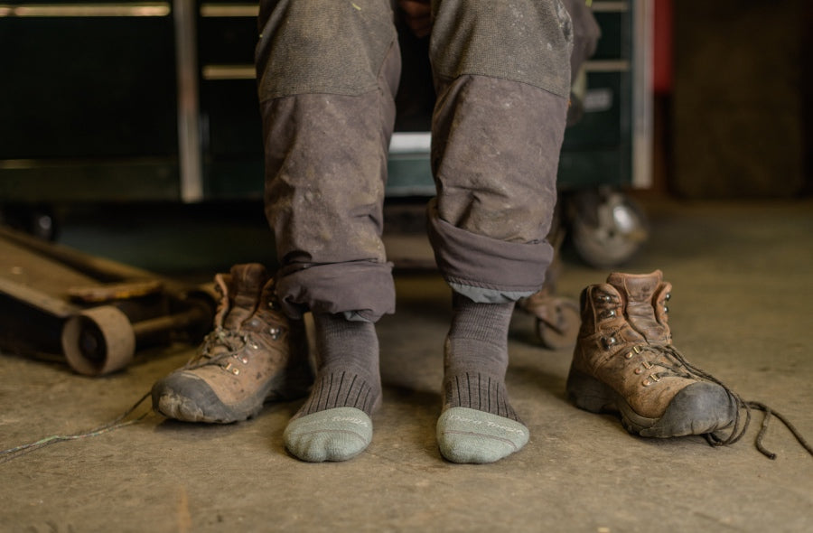 A person's feet in Darn Tough work socks with safety toe boots nearby, about to put on