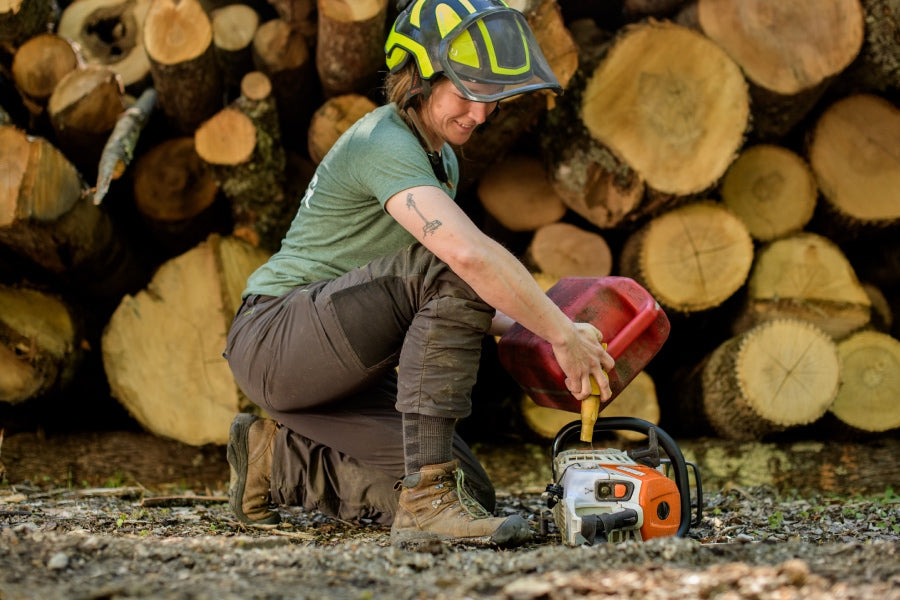 A person with a chainsaw wearing darn tough work socks and steel toe boots by a log stack