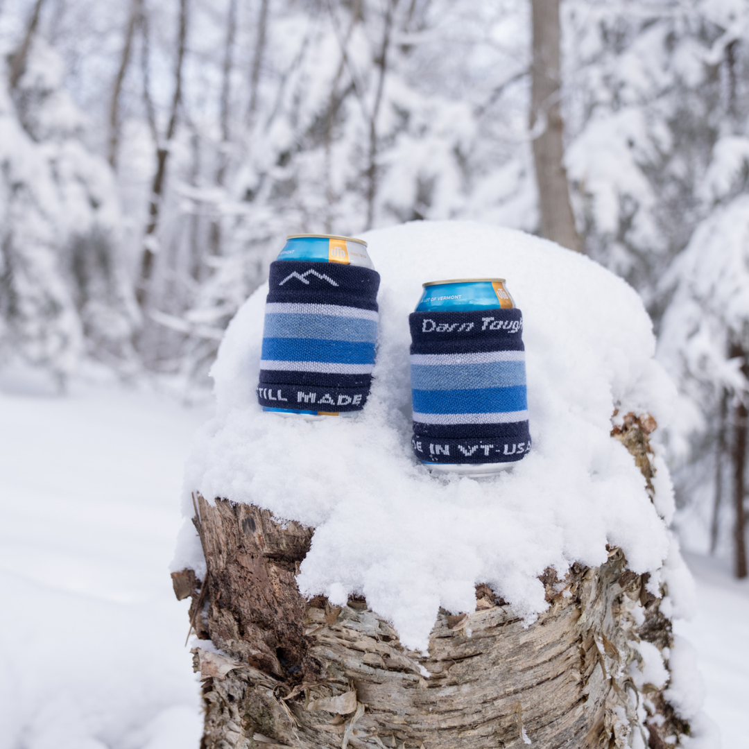 two cans sitting on a snow covered tree stump wrapped in the Via feratta koozies