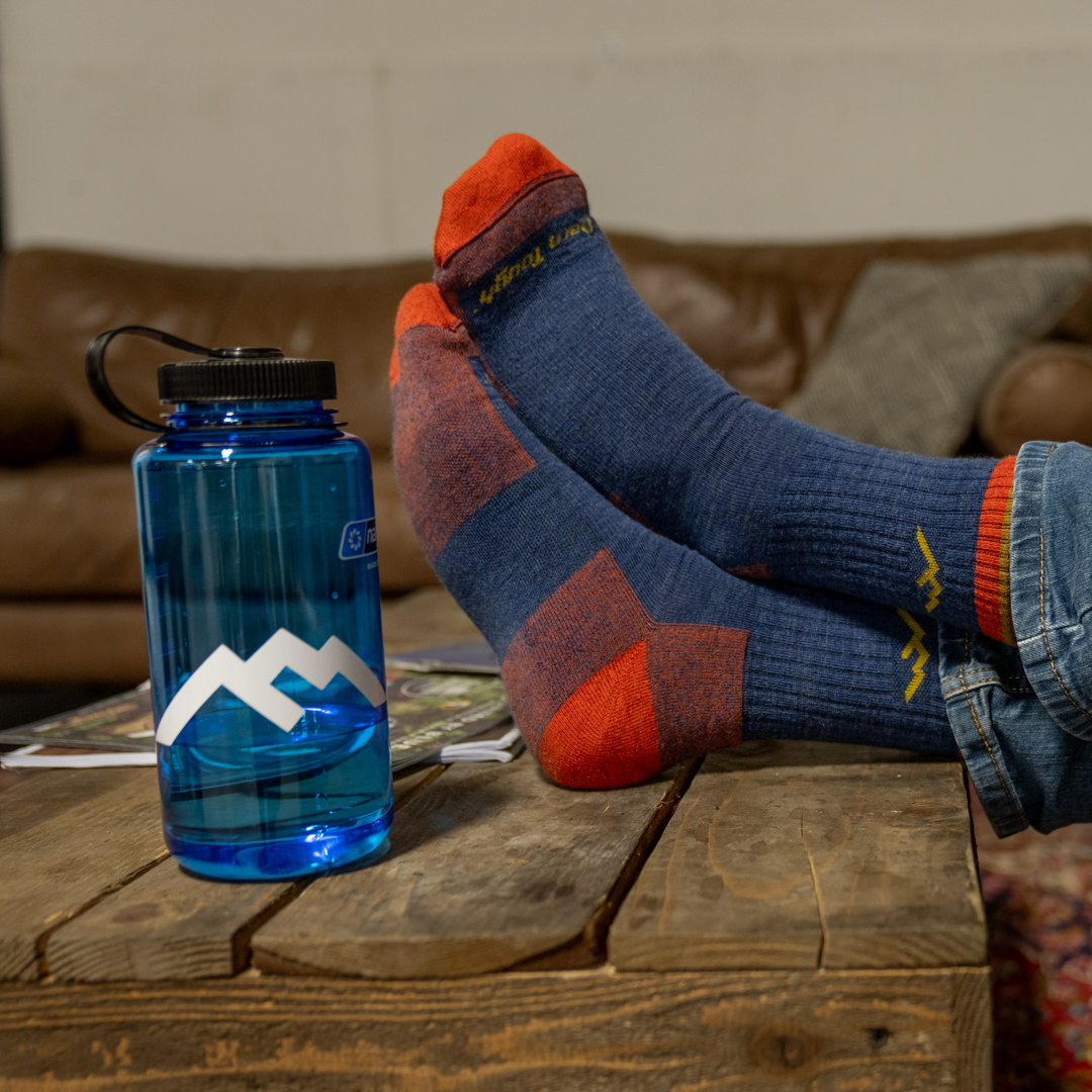 White Darn Tough mountain logo sticker on blue water bottle on wooden table with model's feet in Darn Tough socks propped next to it