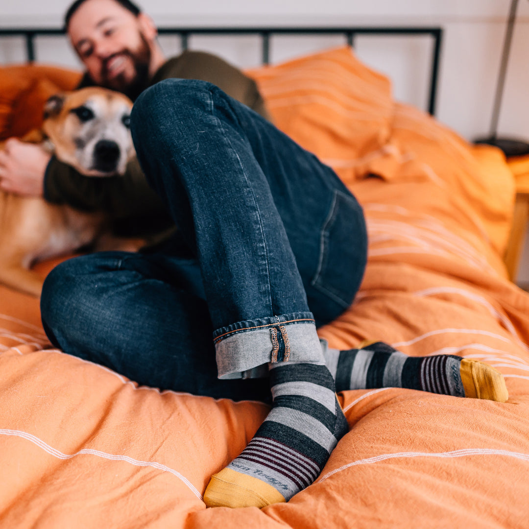 Full shot of model laying in orange bed with their dog wearing the men's oxford crew lifestyle sock in gray
