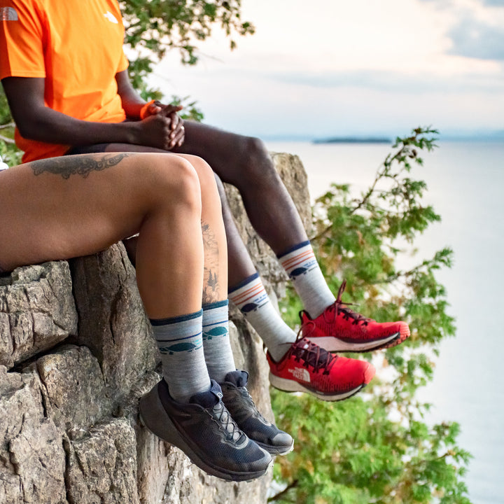 Lifestyle shot of 2 models wearing Pacer socks. The woman is wearing hers in Gray, sitting on a ledge above Lake Champlain.