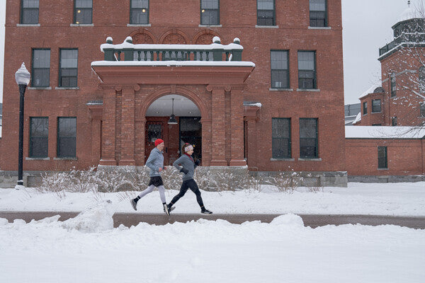 Image of two runners in mid stride with brick building and snow in the background.