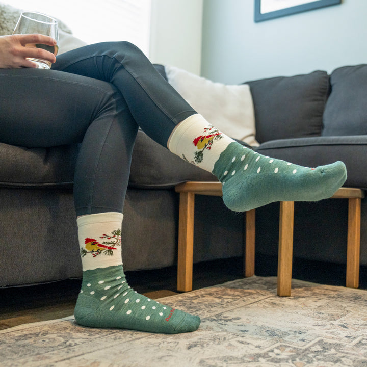 Model sitting on couch wearing the 6209 Wildlife crew sock, featuring green body with white dots and cardinal on the ankle