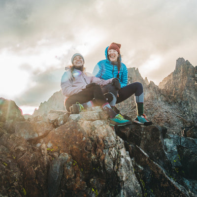 Image of 2 women, laughing, sitting on a rocky mountaintop and one is wearing Women's Bear Town Hiking sock in Purple, Lifestyle Image