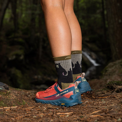 Close up lifestyle shot of model wearing Beartown socks in Oatmeal and orange trail runners, walking in the dimly-lit woods.