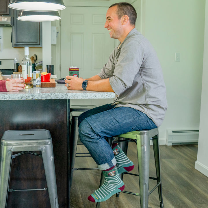 A man sitting at a kitchen Island wearing the 6211 wildlife socks that feature white/green stripes and red heel/toe.