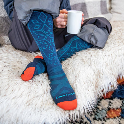 Close up shot of woman sitting on a pelt while drinking a cup of coffee in her Traverse socks in Eclipse, and snowpants.