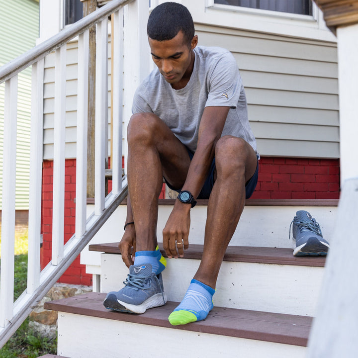 Full body shot of model sitting on steps putting on his sneakers wearing the men's no show tab running sock in surf blue