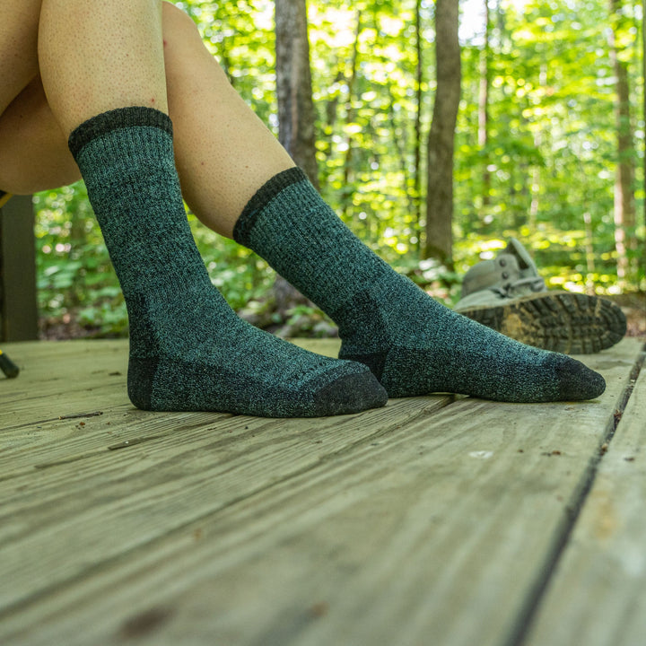 Image of a woman's feet on a wooden porch in the woods, hiking boots in the background, wearing Women's Nomad Boot Midweight Hiking Socks in Aqua, Lifestyle Image