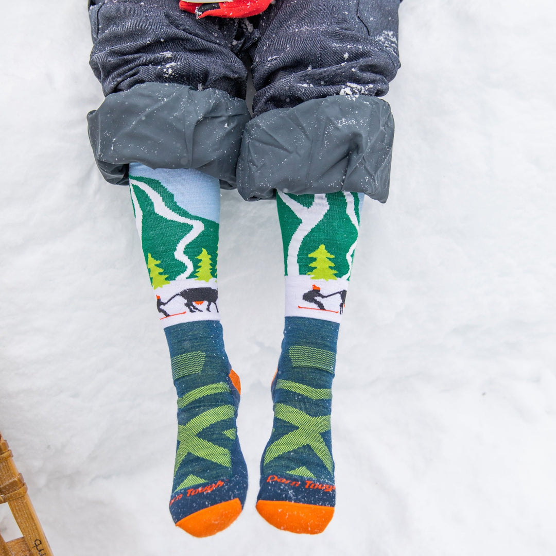 Overhead close up shot of model sitting in the snow wearing the kids' pow cow over-the-calf snow sock in green