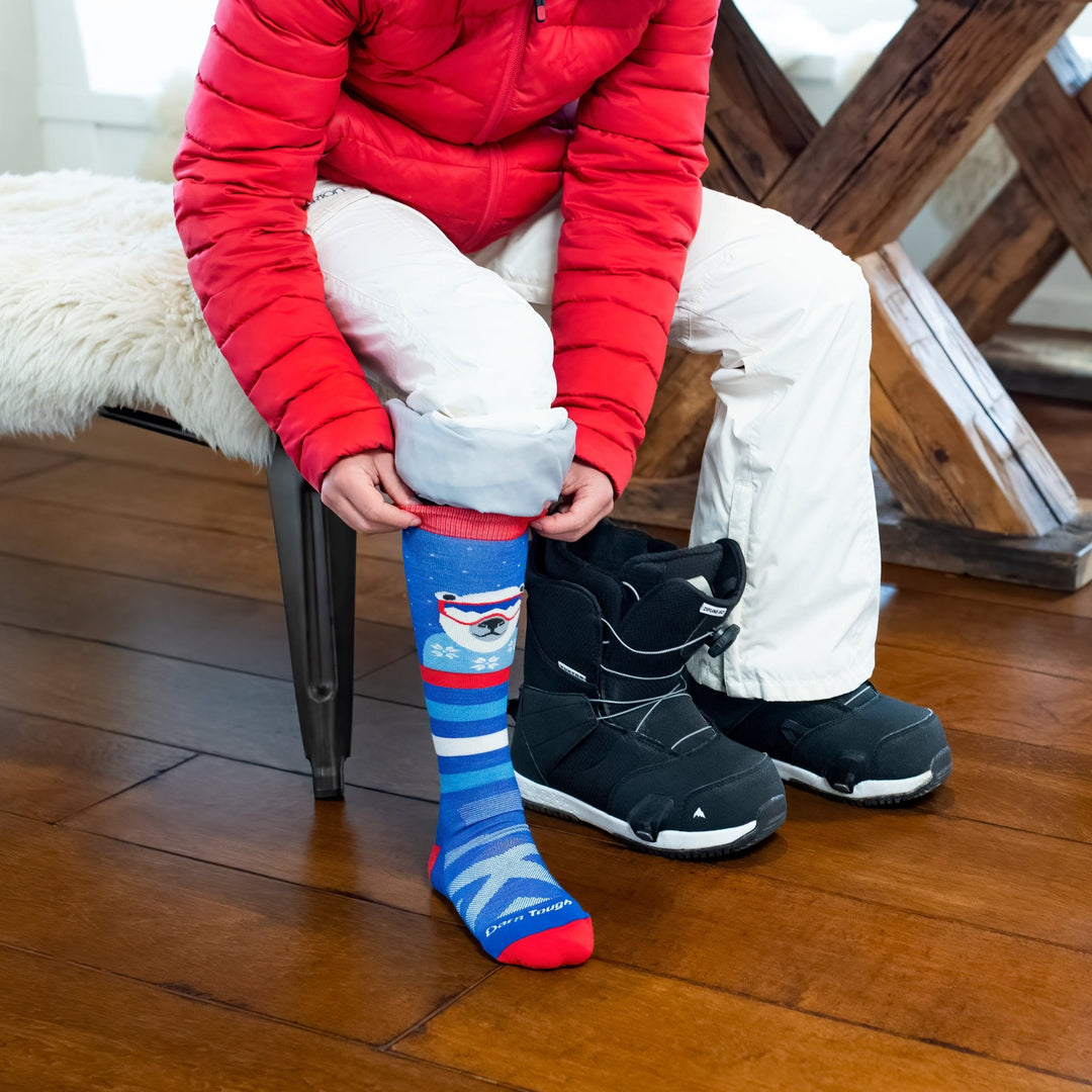 Side shot of model putting on the kids' polar patroller over-the-calf snow sock with black snowboard boots