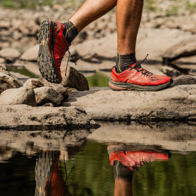 Side shot of model walking on river rocks wearing the men's quarter hiking sock in taupe with red hiking shoes on
