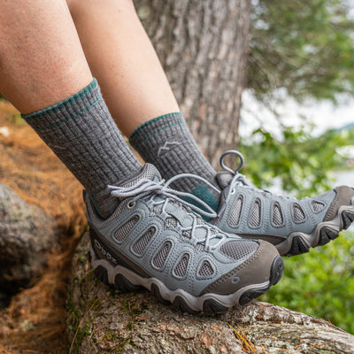 Close up shot of model sitting on a tree stump wearing women's micro crew hiking sock in slate with light blue hiking shoes on