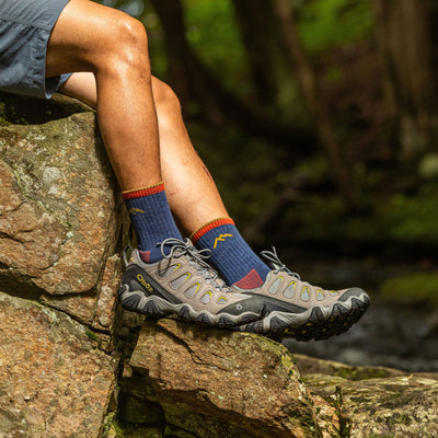 Side shot of model sitting on a river rock wearing the men's micro crew hiking sock in denim with brown hiking shoes
