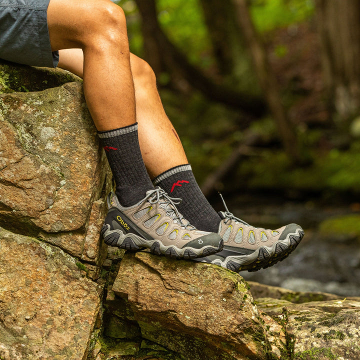Side shot of model sitting on river rocks wearing men's micro crew hiking sock in black with brown hiking shoes on