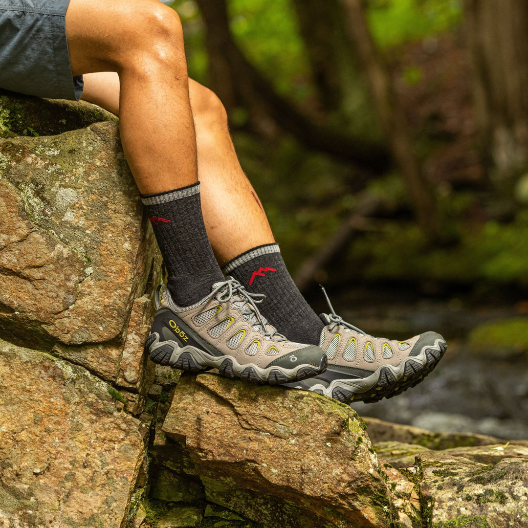 Model sitting on a rive rock wearing men's micro crew hiking sock in black with brown and gray hiking shoes 