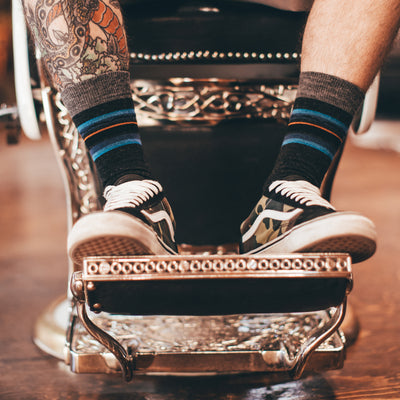 Man sitting in a barber shop chair, wearing sneakers and Whetstone Crew Lightweight Lifestyle socks in Charcoal, Lifestyle Image