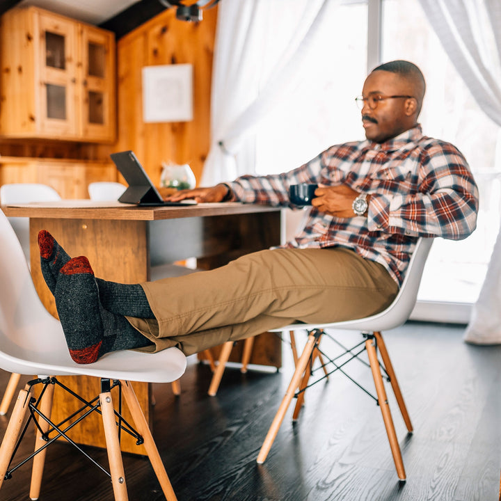 Full shot of model sitting at a desk with his feet up on a white chair wearing men's nomad boot hiking sock in pewter gray