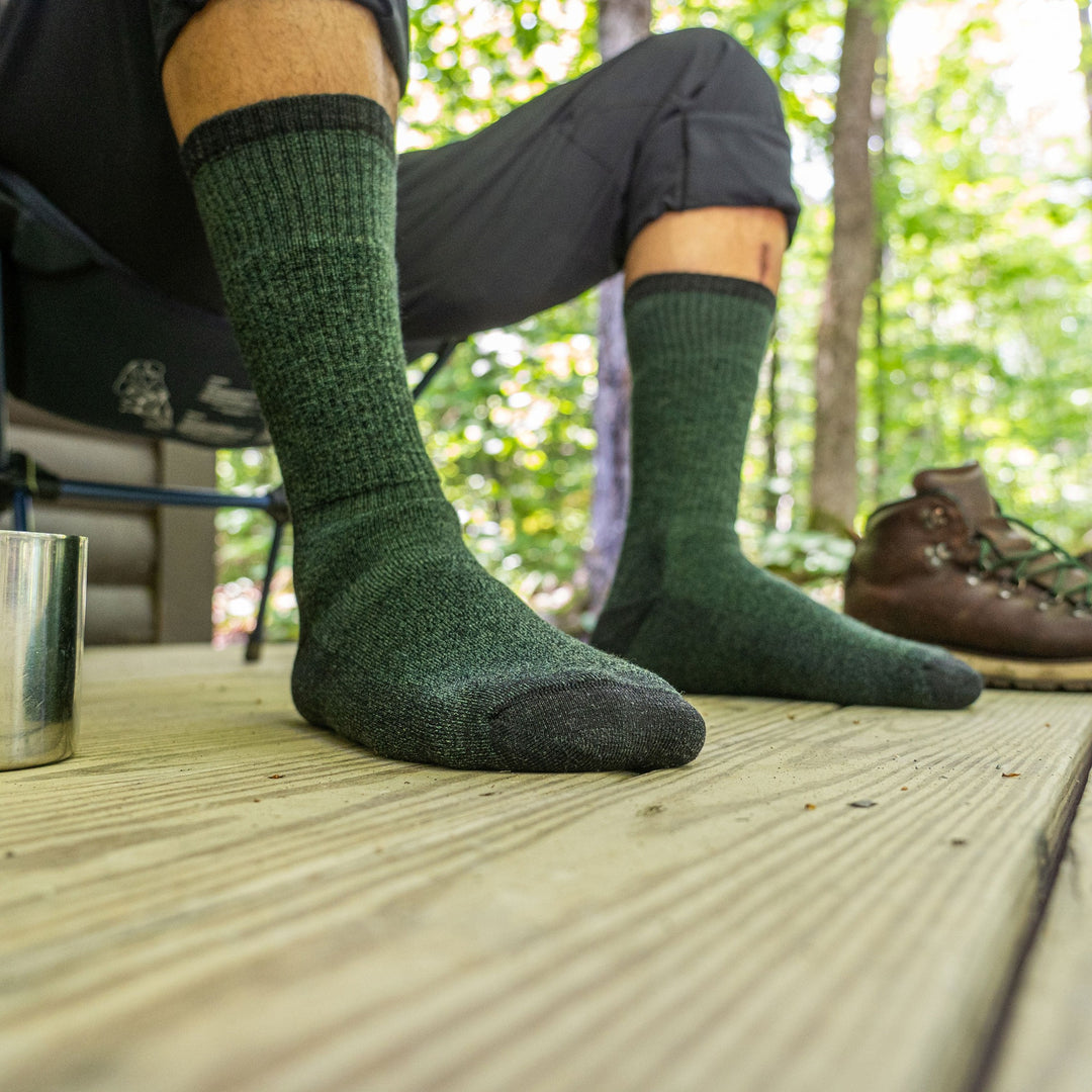 Close up lifestyle image of a man's feet while sitting in a camping chair on a porch, hiking boots nearby wearing Nomad Boot Midweight Hiking Socks in Moss, Lifestyle Image