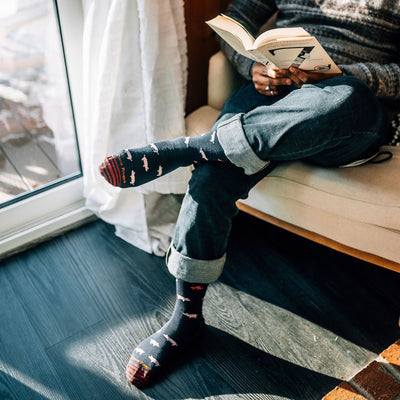Half shot of model sitting on a white chair inside reading a book wearing men's truffle hog crew lifestyle sock in navy blue