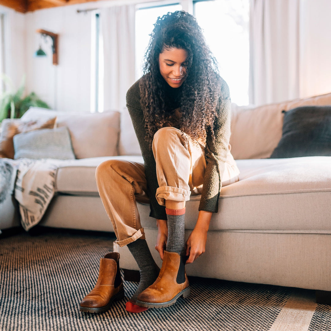Full shot of model sitting on tan couch wearing women's nomad boot hiking sock in brown while putting on brown short boots