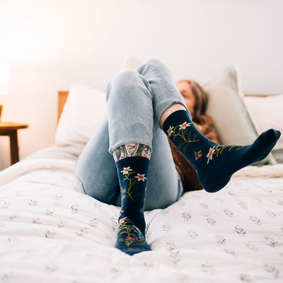 Full shot of model laying in a white bed wearing women's fable crew lifestyle sock in denim blue