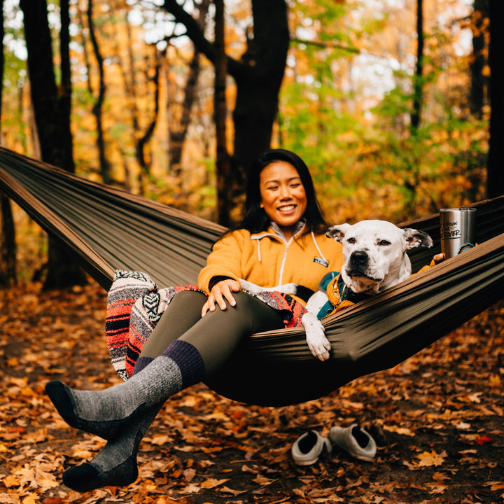 Lifestyle shot of woman smiling with her dog in her hammock. It is fall in Vermont and she's wearing her Scout socks in Plum.
