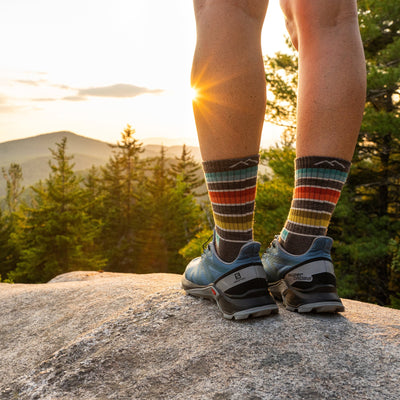 A woman stands on a rock wearing sneakers and Women's Decade Stripe Micro Crew Midweight Hiking Socks in Taupe, looking out over a forest and mountain range as the sun sets, Lifestyle Image